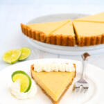 Slice of key lime tart on plate with whipped cream and lime wheel garnish. There is a silver fork on the side of the pie.