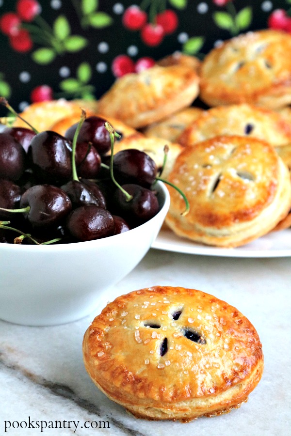 cherry pie with bowl of cherries and plate of hand pies in background