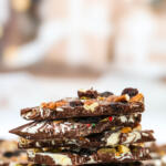 Pieces of fruit and nut chocolate bark stacked on top of each other, sitting on top of a piece of brown parchment paper.
