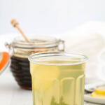 Glass of cold remedy tea with lemon and ginger in front a pot of honey.
