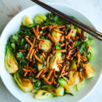 Sautéed baby bok choy with gochujang mushrooms in a white bowl. There is a pair of chopsticks laying across the top of the bowl.