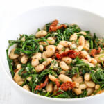 White beans with arugula and sun-dried tomatoes in a white bowl.