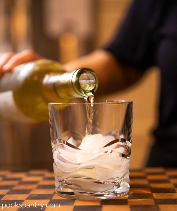 Pouring white wine into old-fashioned glass sitting on wooden board to make spritzer drink.