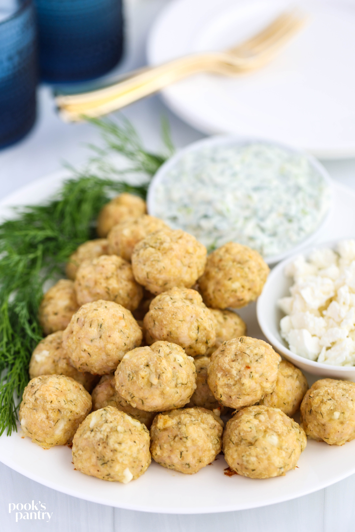 Chicken meatballs stacked on a white plate with fresh dill, and small bowls of feta and tzatziki.