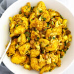 Curried cauliflower with golden raisins and ginger in a white bowl with a blue towel on the side.