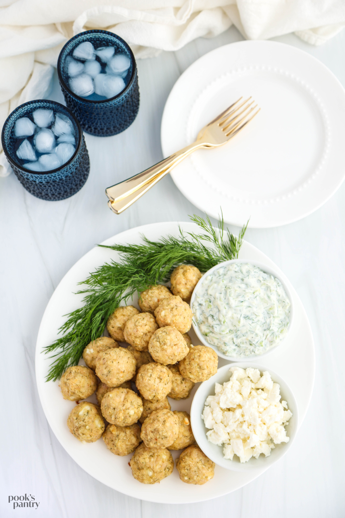 Overhead photo of chicken meatballs, plates with forks on top, and two glasses of ice water, on a white background.