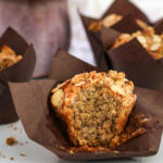 Banana muffin in brown muffin paper with crumbles of streusel topping in front.