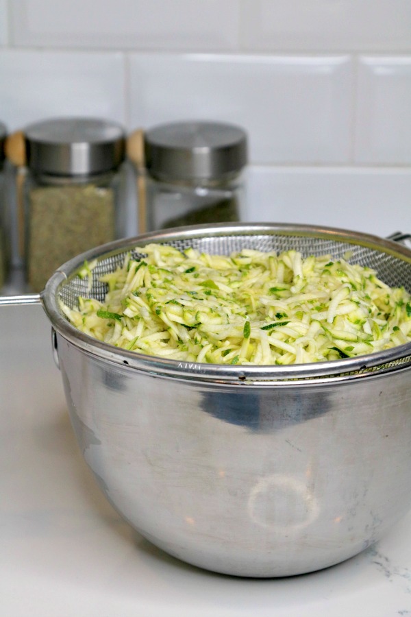 Grated zucchini in fine mesh colander inside of stainless steel mixing bowl on countertop.
