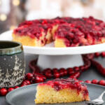 Cranberry upside down cake on black plate, with the whole cake on a stand in the background.