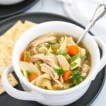 Classic chicken noodle soup in a white soup crock on a black plate.