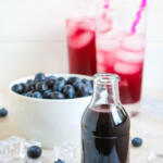 Blueberry syrup for drinks in a small glass bottle in front of two glasses of blueberry soda water.