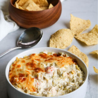 Langostino artichoke dip in straight-sided white bowl with tortilla chips in the background.