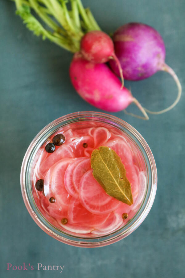Overhead view of glass canning jar with radish and spices, raw radishes in background on gray slate.