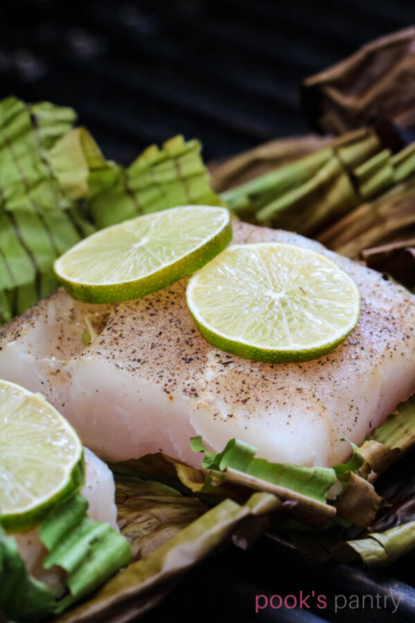 Grilled corvina fillet on banana leaves on grill grates with limes wheels on top of fish.