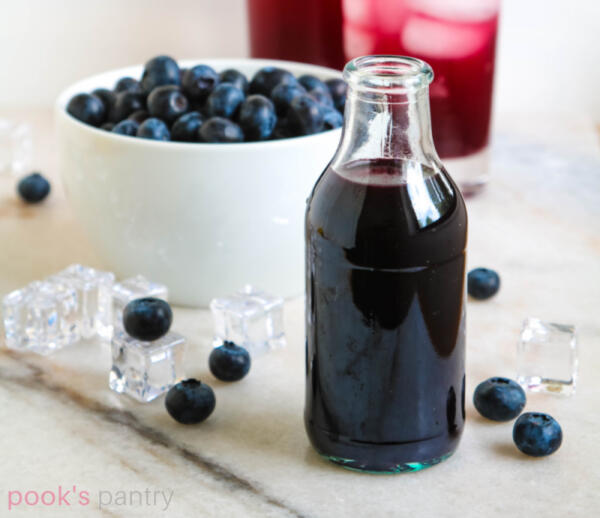 Blueberry syrup for lemonade in small glass bottle with blueberries scattered around white marble background.