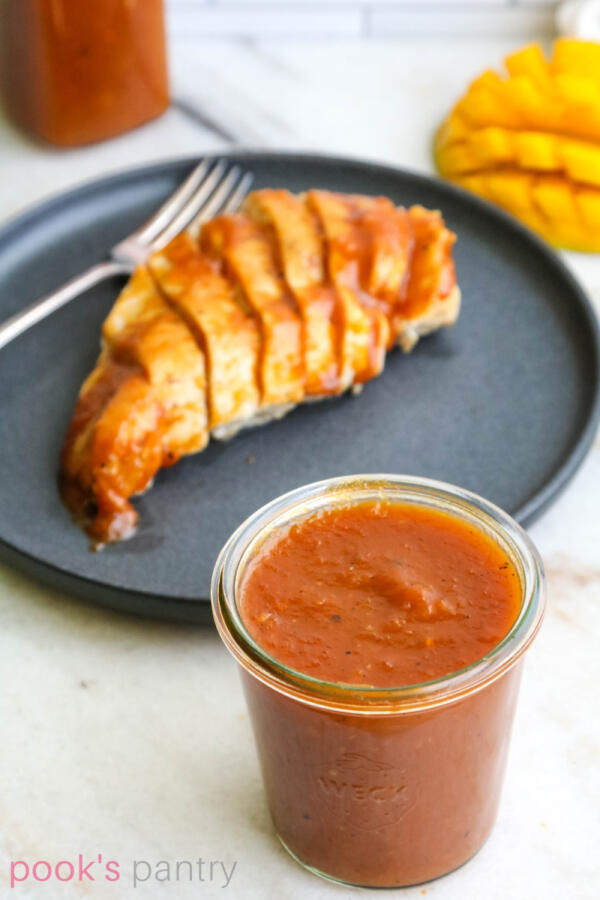 Pineapple mango BBQ sauce in glass jar with chicken breast on plate in background.