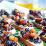 Apple chutney crostini on a white platter with glasses of wine in the background.