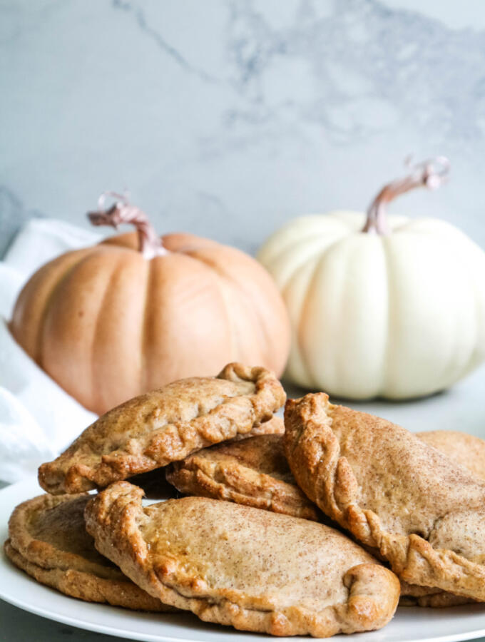 Pumpkin empanadas on white plate on gray marble backdrop with pumpkins in the background.