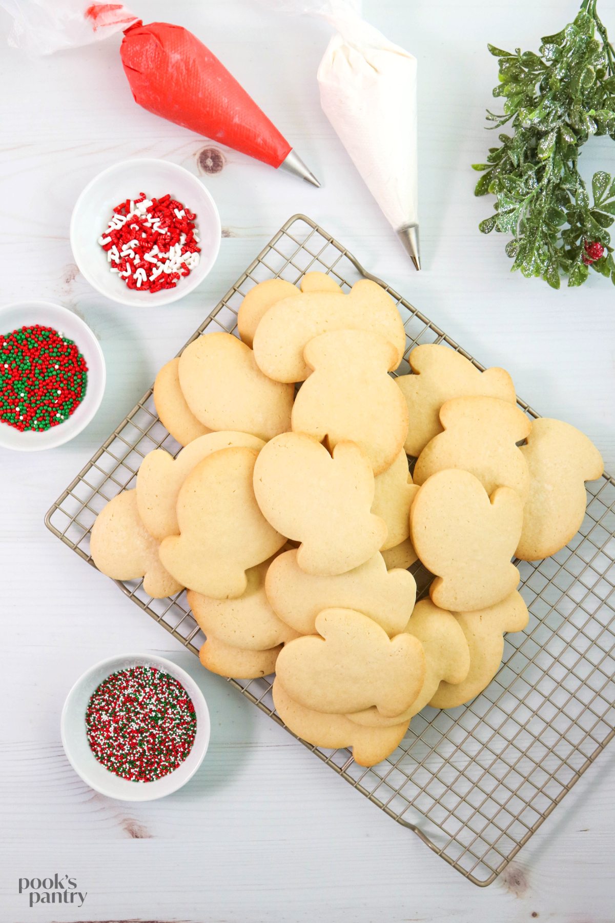 Classic sugar cookies piled on to a metal cooling rack, with piping bags full of icing, and small bowls of sprinkles.