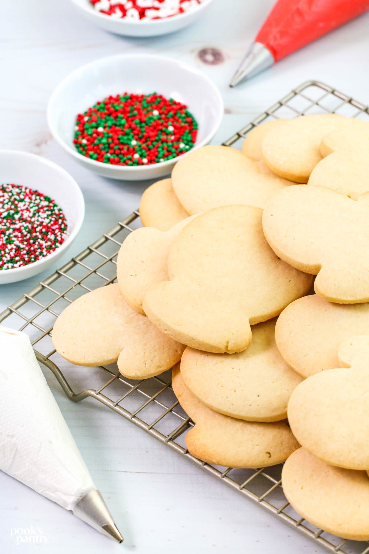 Classic sugar cookies piled on a metal cooling rack. There are small bowls of sprinkles and piping bags filled with icing in the background.