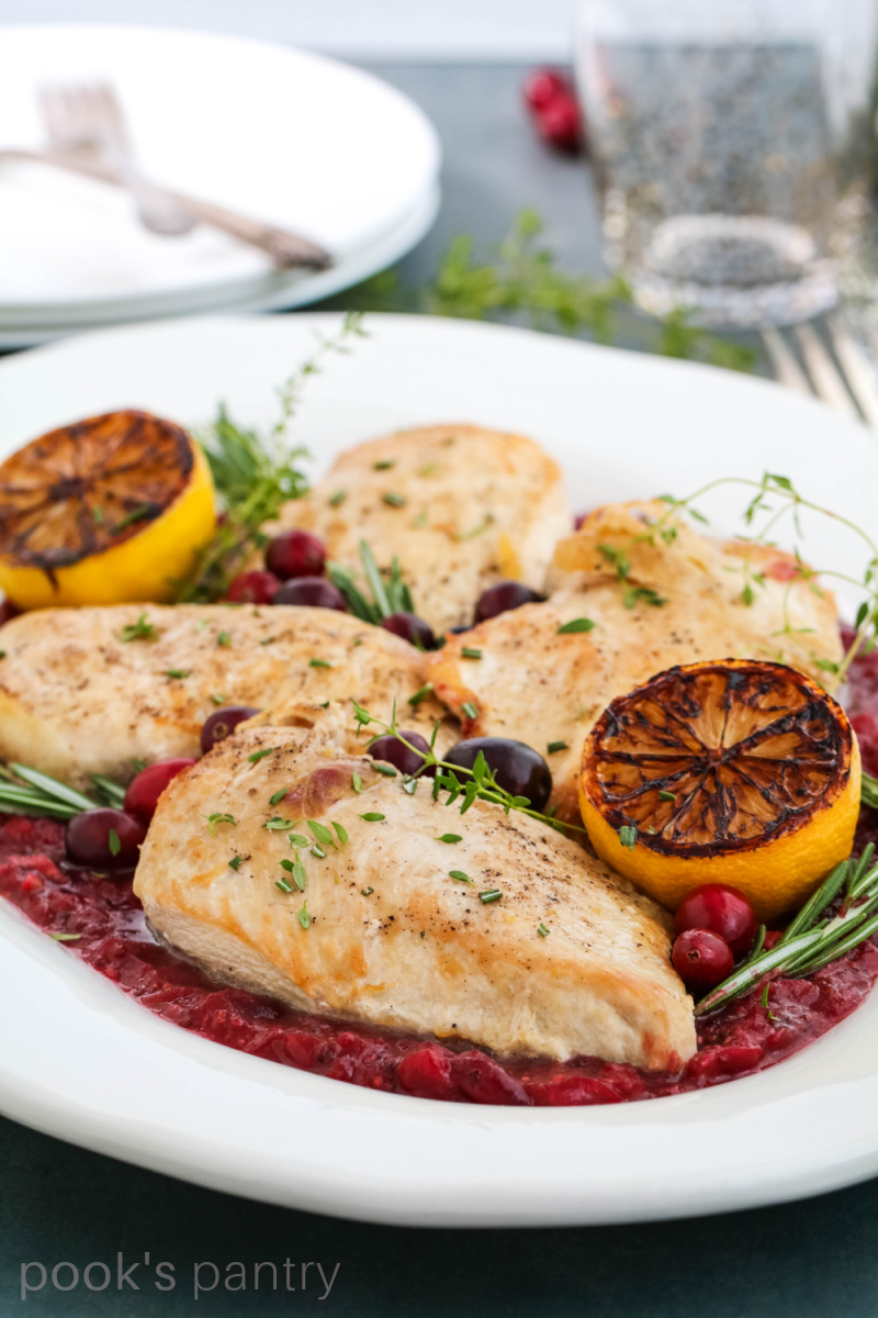 Cranberry chicken on a white platter with charred lemons and rosemary on the side.