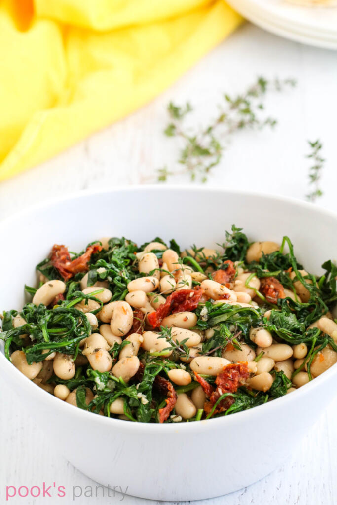 Beans with sun dried tomatoes and arugula in white bowl.