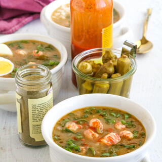 Langostino gumbo in white soup crocks with pickled okra and hot sauce in the background.
