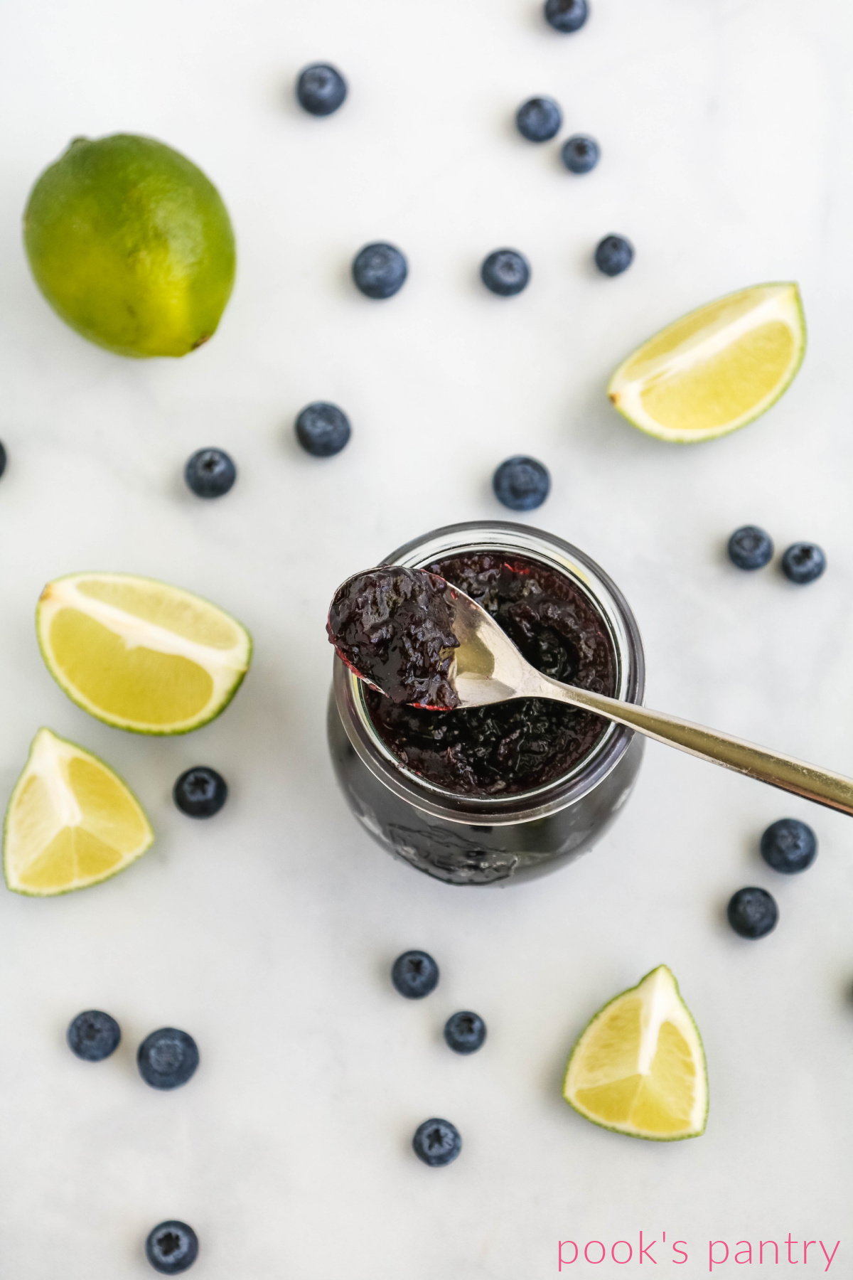 Blueberry lime jam on spoon with blueberries and limes in the background.