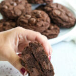 Hand holding a chocolate chocolate chip cookie cut in half.