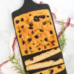 Rosemary focaccia with black olives on a black walnut cutting board, with sprigs of rosemary on the sides.