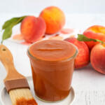 Peach BBQ sauce in jar on a small white plate with a pastry brush on the side. There are whole peaches in the background.