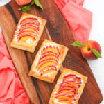 Fresh peach puff pastry tarts on brown wooden board with whole peaches on the side.