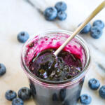 Blueberry bourbon jam recipe in a glass canning jar with a small silver spoon in the jar. There are blueberries scattered around the jar.