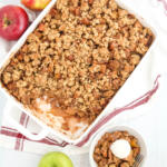 Easy apple crisp recipe in white casserole dish with 3 different colored apples around the pan. There is a small bowl of apple crisp sitting in front of the pan.