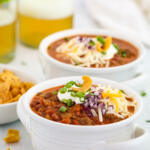 Beef and bean chili in white soup crocks with small glasses of beer in the background.