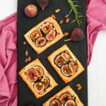 Four savory fig tarts on a black wooden board with figs placed beside them. There is a purple linen towel under the board, with pistachios and a sprig of rosemary.