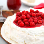 Pavlova with whipped cream and raspberry on white platter with jar of raspberry sauce in the background.
