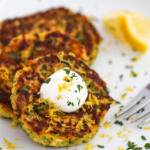 Zucchini fritters recipe with lemon zest and herbs on a white plate with lemon wedges and sour cream.