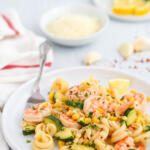 Shrimp and zucchini pasta with corn on a white plate with cheese, garlic, lemon and herbs in the background.
