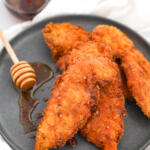 Hot honey chicken tenders on a black plate with a honey dipper sitting in a puddle of honey on the side.