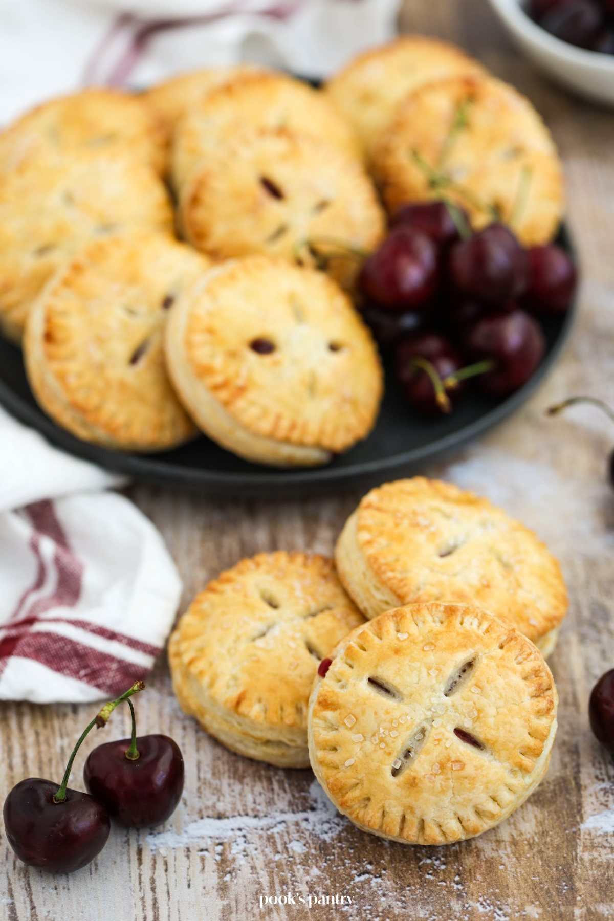 Cherry hand pies on a table with fresh cherries and a kitchen towel beside them.