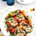 Baked corvina recipe with tomatoes, olives and capers on a white platter with blue drinking glasses in the background.
