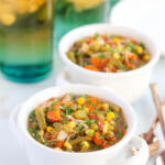 CrockPot chicken and vegetable soup on white soup crocks with teal and yellow glasses in the background.