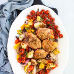Baked chicken thighs and vegetables on a white oval platter sitting on a black wooden board.