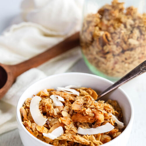 Coconut granola recipe is a white bowl with a spoon. There is a clear glass jar filled with granola in the background.