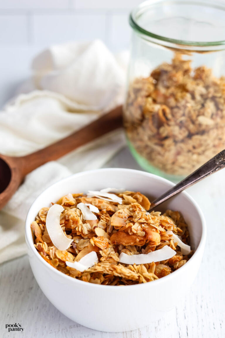 Coconut granola recipe is a white bowl with a spoon. There is a clear glass jar filled with granola in the background.