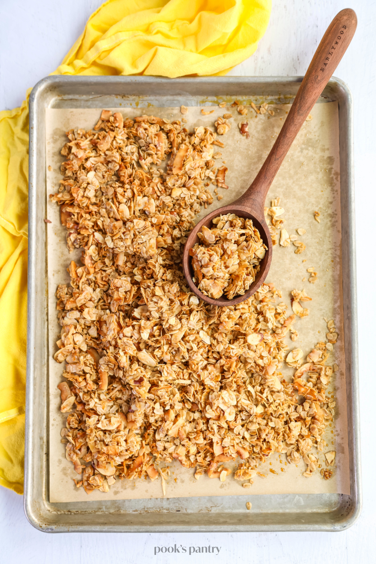 A wooden ladle filled with granola on a sheet pan full of granola, with a yellow tea towel on the left side.