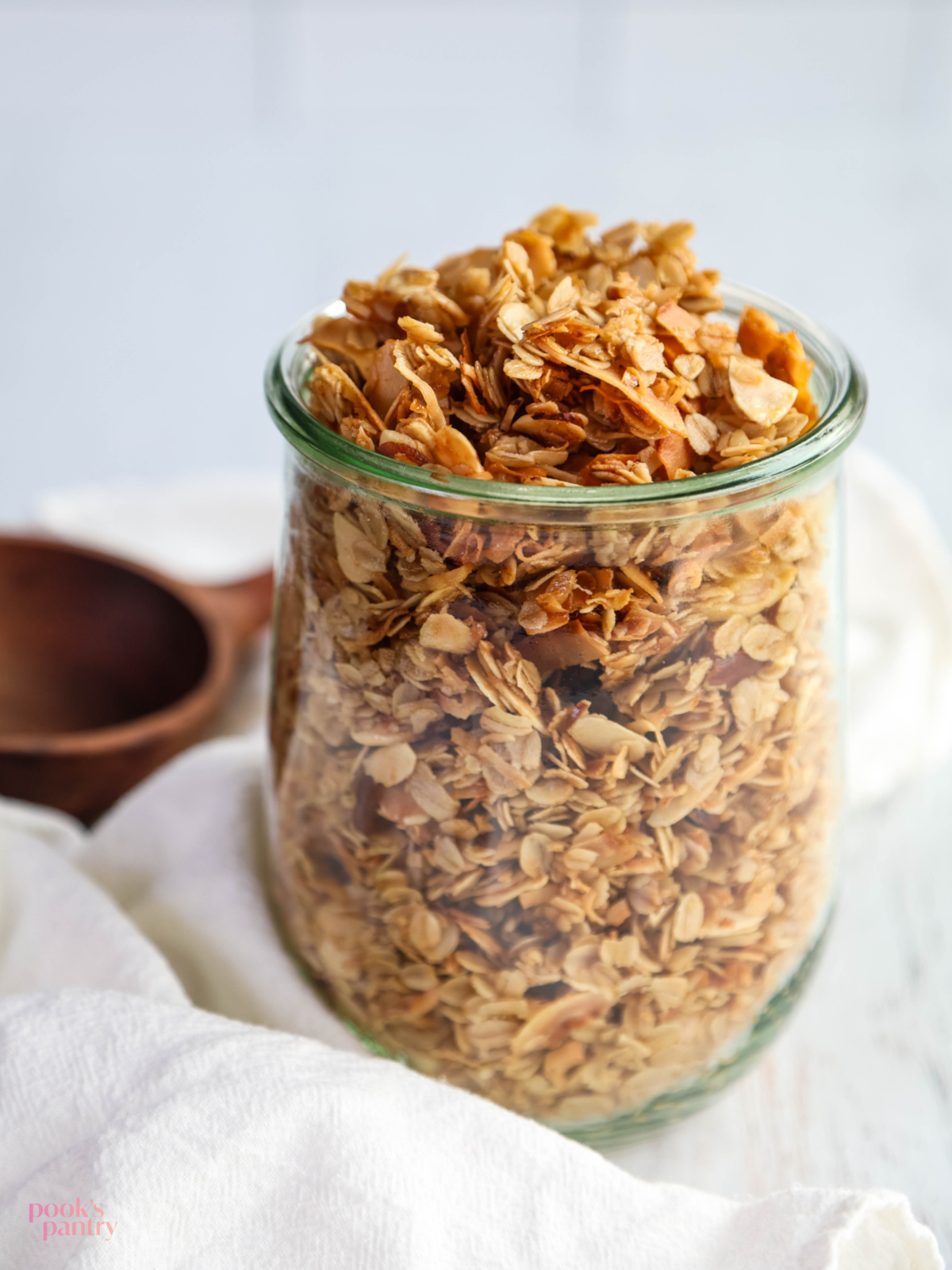 Coconut granola in a glass jar next to a white tea towel with a wooden ladle in the background.