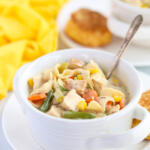 Creamy chicken noodle soup in a white soup crock bowl with handles and an antique spoon in the bowl.