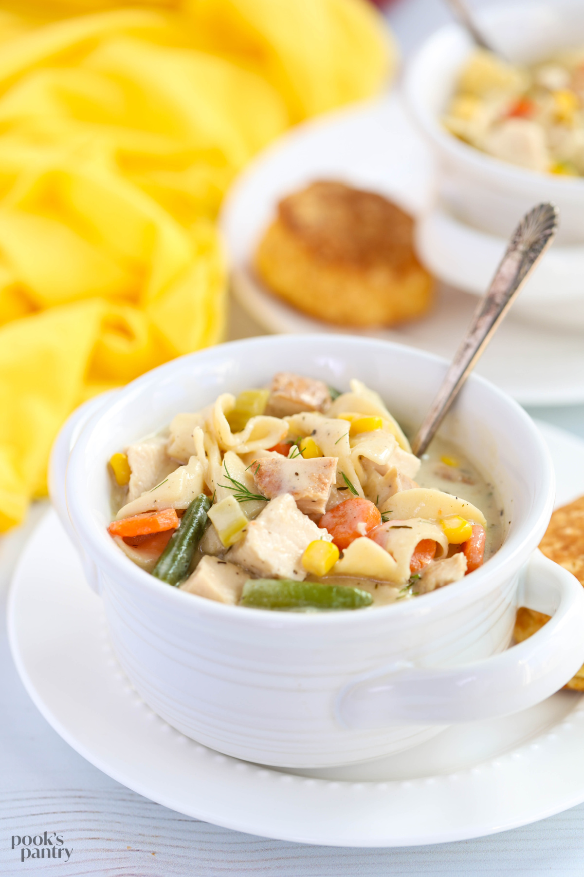 Creamy chicken noodle soup in a white soup crock bowl with handles and an antique spoon in the bowl.